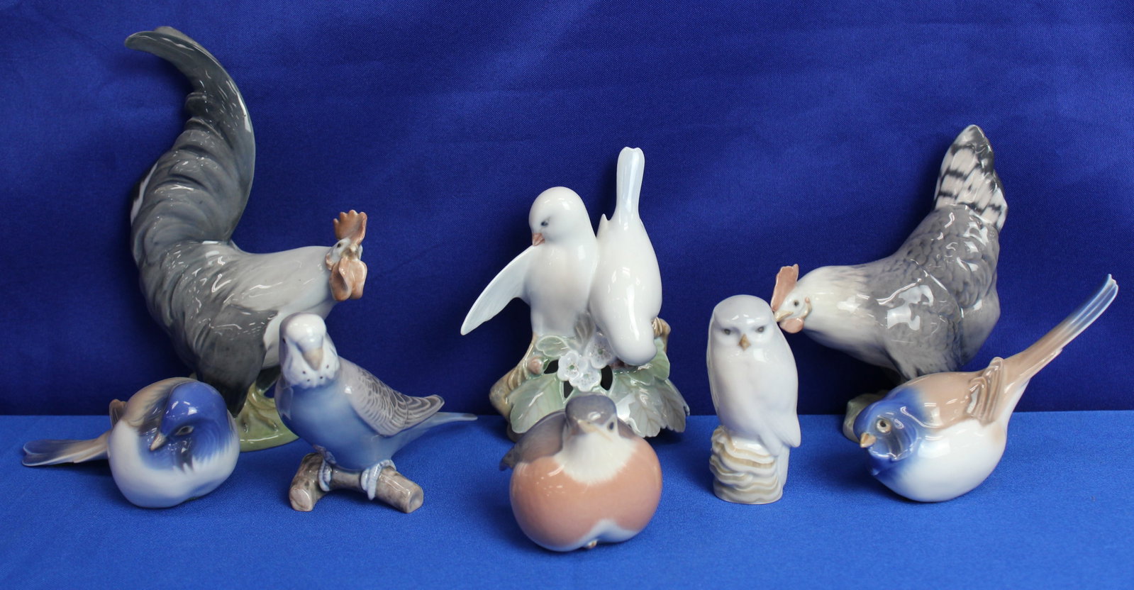 Royal Copenhagen Bird Figurines: Collection of Royal Copenhagen bird figurines featuring the fledgling robin, B&G parakeet, white owl, 2 titmouse birds, 2 roosters, and the two doves figurine. All the figurines have royal Copenhagen