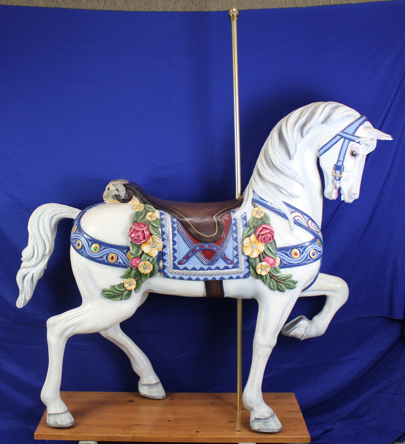 Large Wood Carousel Horse: White wood carousel horse with blue saddle adorned with flowers and plastic blue, green, and red gems. The back of the saddle has the head of a goat and the horse has a metal gold tone pole going thro