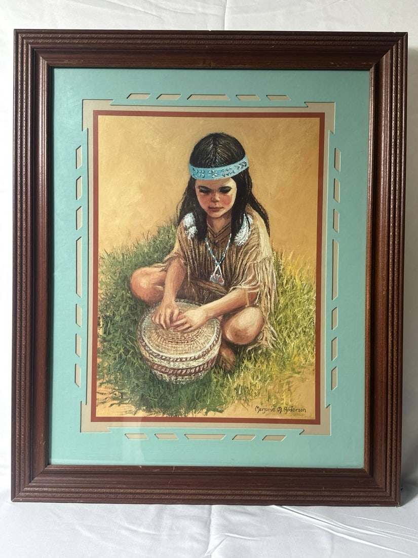 Marjorie M Anderson Framed Native American Child Art 19x23x1 in: This captivating artwork by Marjorie M. Anderson features a tender depiction of a young Native American child, sat thoughtfully on grass. Playing gently with a drum, the child wears a detailed traditi