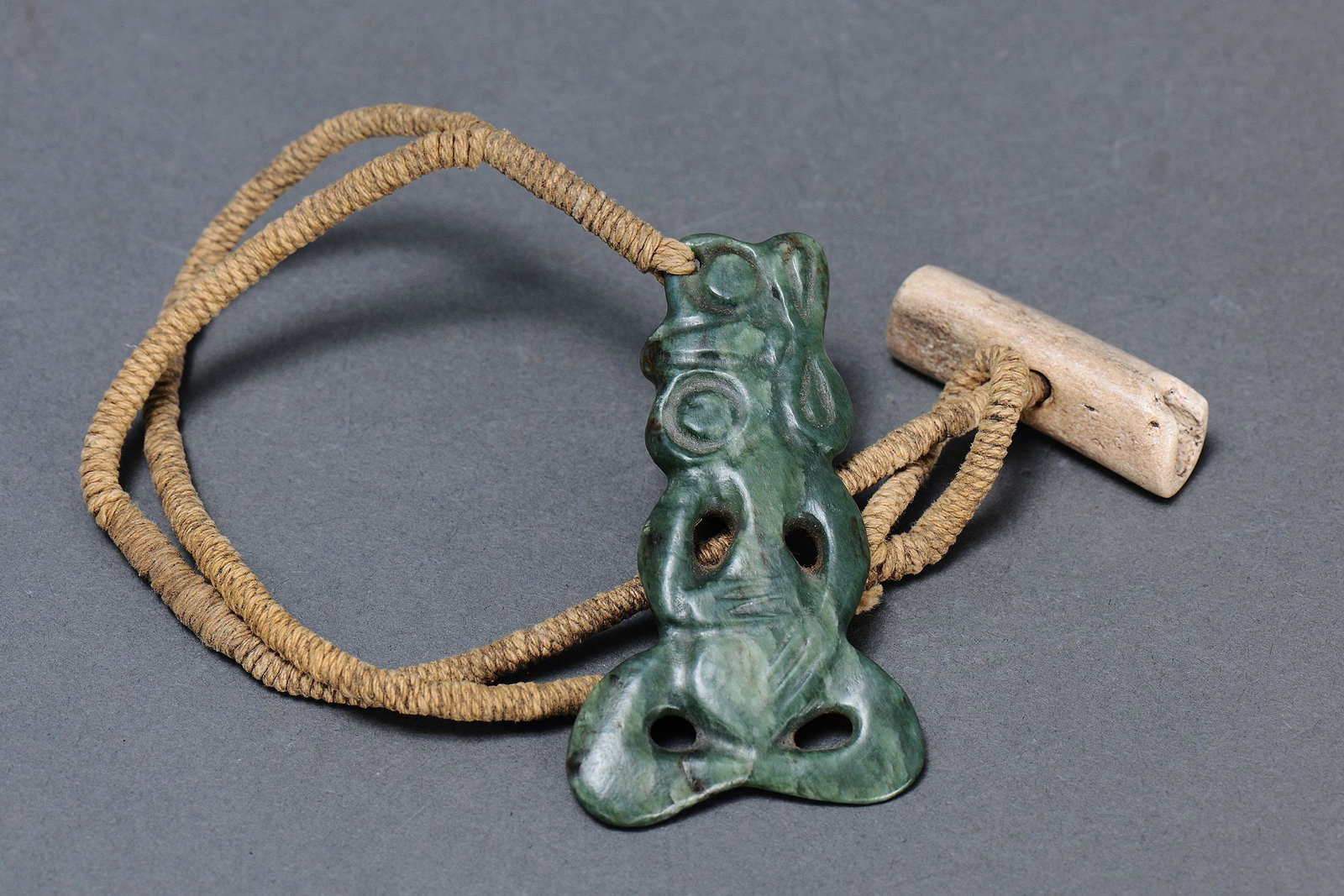MAORI ‘HEI TIKI' POUNAMU (GREENSTONE) PENDANT, NEW ZEALAND: Maori ‘Hei Tiki' Pendant, New Zealand. Carved and engraved Pounamu (Greenstone) attached to Flax fibre Cord with Bone Toggle. Hei means neck ornament and tikki means human. It was worn on the br