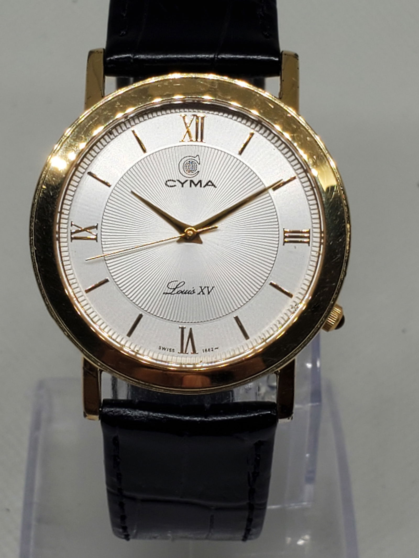 Cyma Louis XV Gold Plated Unisex Watch, 1990s, Working: Elevate your collection with this elegant Cyma Louis XV dress watch, a perfect combination of Swiss refinement and timeless prestige. The 33mm gold tone case frames a clean white dial with Roman numer