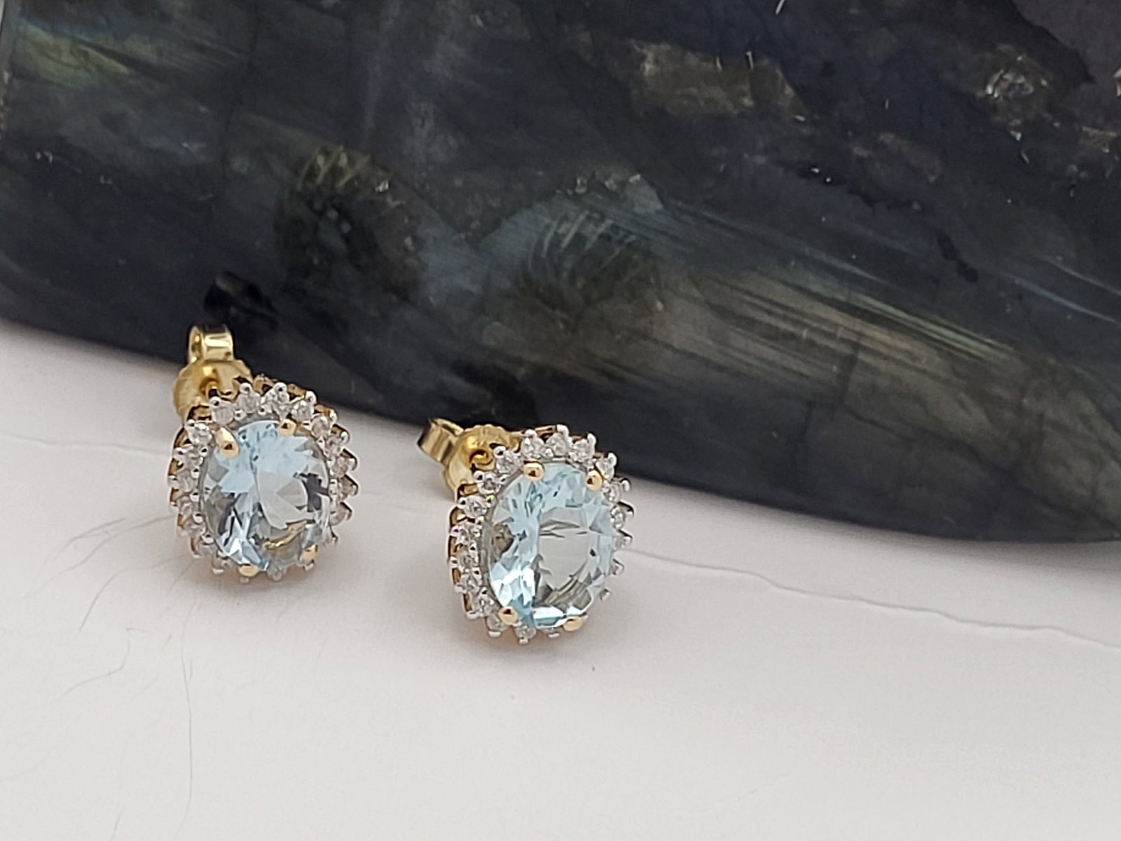 Glacier Blue Aquamarine and Diamond Halo Earrings: Setting: 14K Yellow Gold Main Gemstone: Genuine Aquamarine Adjacent Gemstone: Genuine, Natural Diamond Closure: Posts and Friction Backs Weight: 2.77g Birthstone for: MARCH Aquamarine gemstones can ra