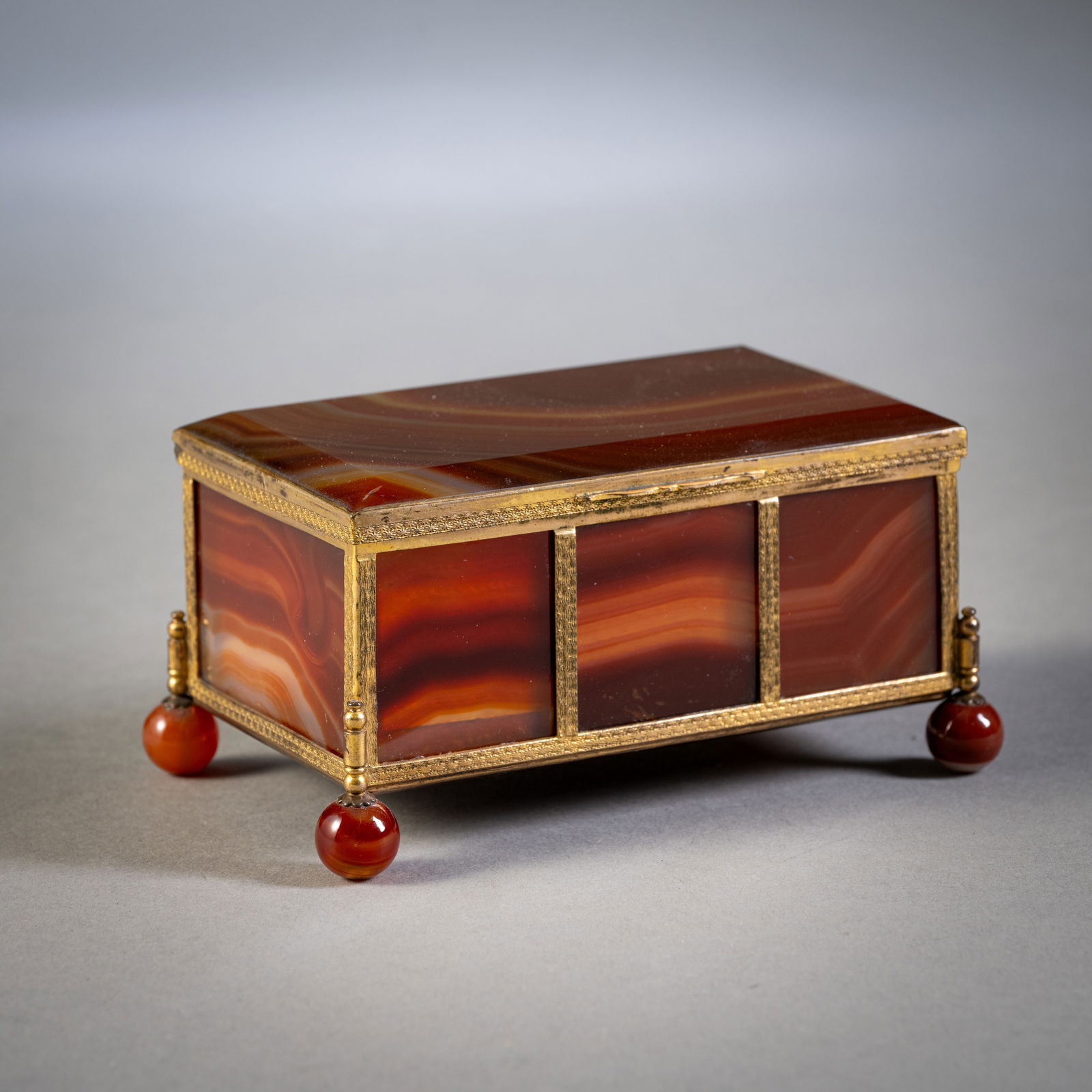 AN AGATE BOX: AN AGATE BOX20th century5,6 x 12,5 x 7,5 cmBrass mounted, on four feet. Minute damage to foot.Achatschatulle20. Jhdt.5,6 x 12,5 x 7,5 cmRechteckig auf 4 Kugelfüßen, Messingmontierung. Minim.