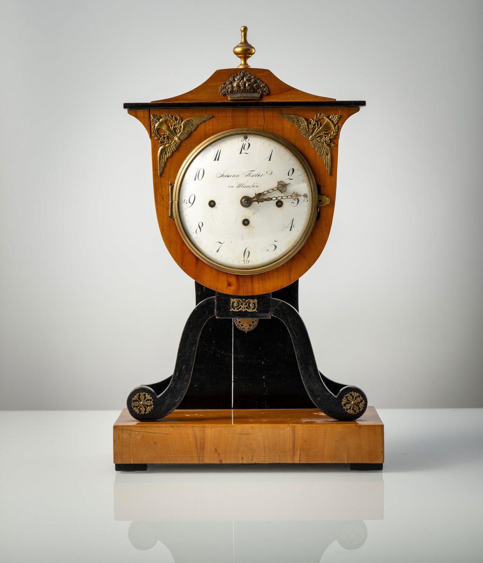 A GERMAN BIEDERMEIER TABLE CLOCK: A GERMAN BIEDERMEIER TABLE CLOCK Munich, circa 1820 H. 53 cm Partially ebonized brass mounted sherrywood case, white enamel dial signed Johann Fischer in München. Movement with 1 day running