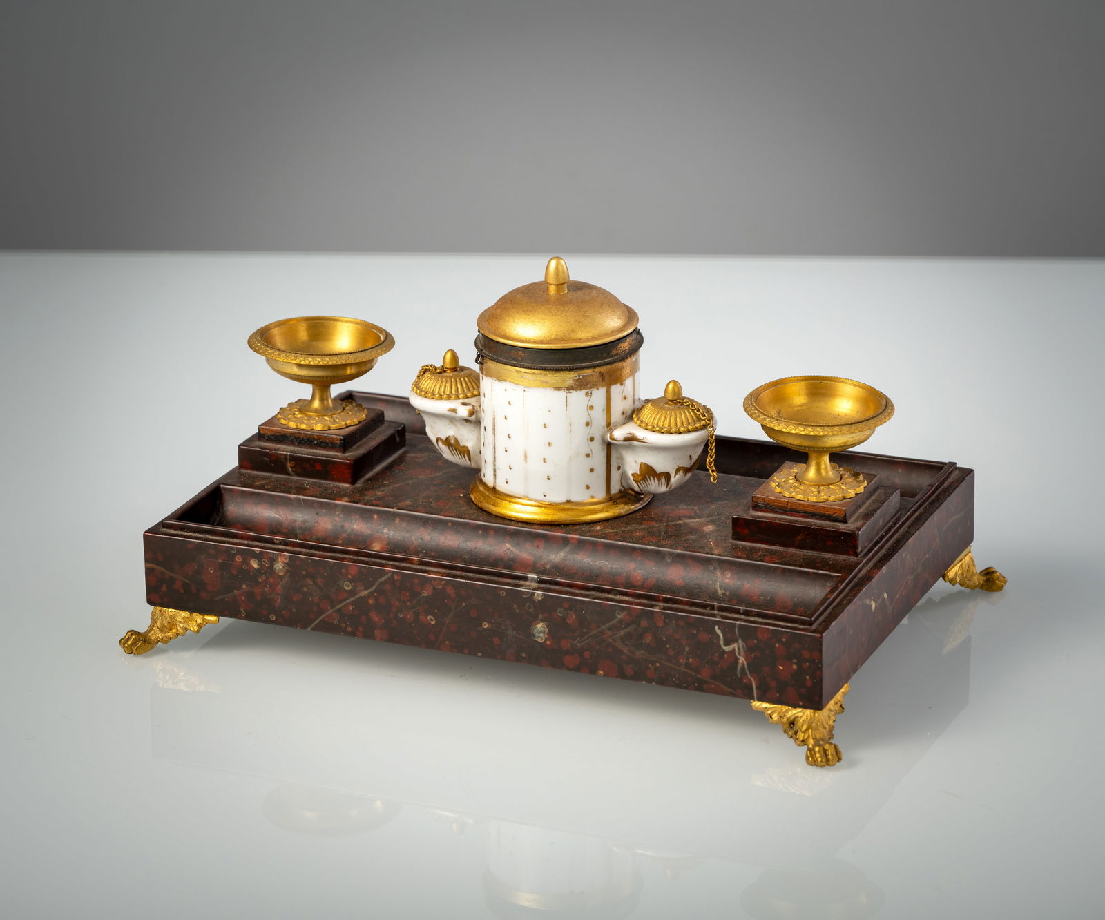 A FRENCH GILT METAL MOUNTED FAUX MARBLE WRITING SET: A FRENCH GILT METAL MOUNTED FAUX MARBLE WRITING SET 1st half 19th century 16 x 28,5 x 19,5 cm Trompe d'Oeil painted wood, gild metal feet and dishes, a central porcelain ink stand with giltwood lid.
