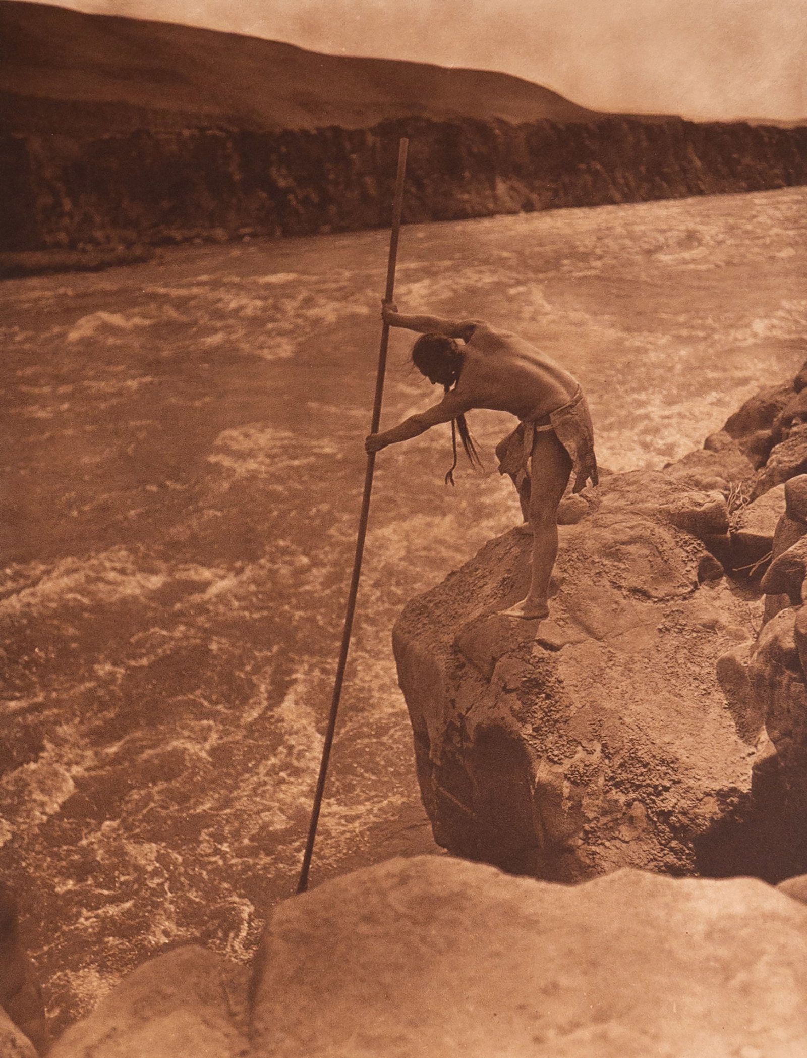 The Fisherman - Wishham: A majestic and atmospheric image capturing a Wishham man fishing from a precarious platform over the Columbia River. With his net extended and body poised in concentration, the fisherman becomes part 