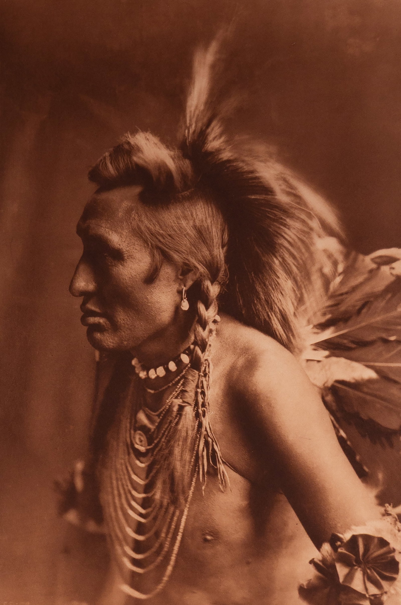 Gambler - Piegan: This striking photograph, taken in 1900, depicts a Piegan Blackfeet man known as the Gambler. Seated in profile against a neutral backdrop, he is dressed in traditional clothing with elaborate beadwor