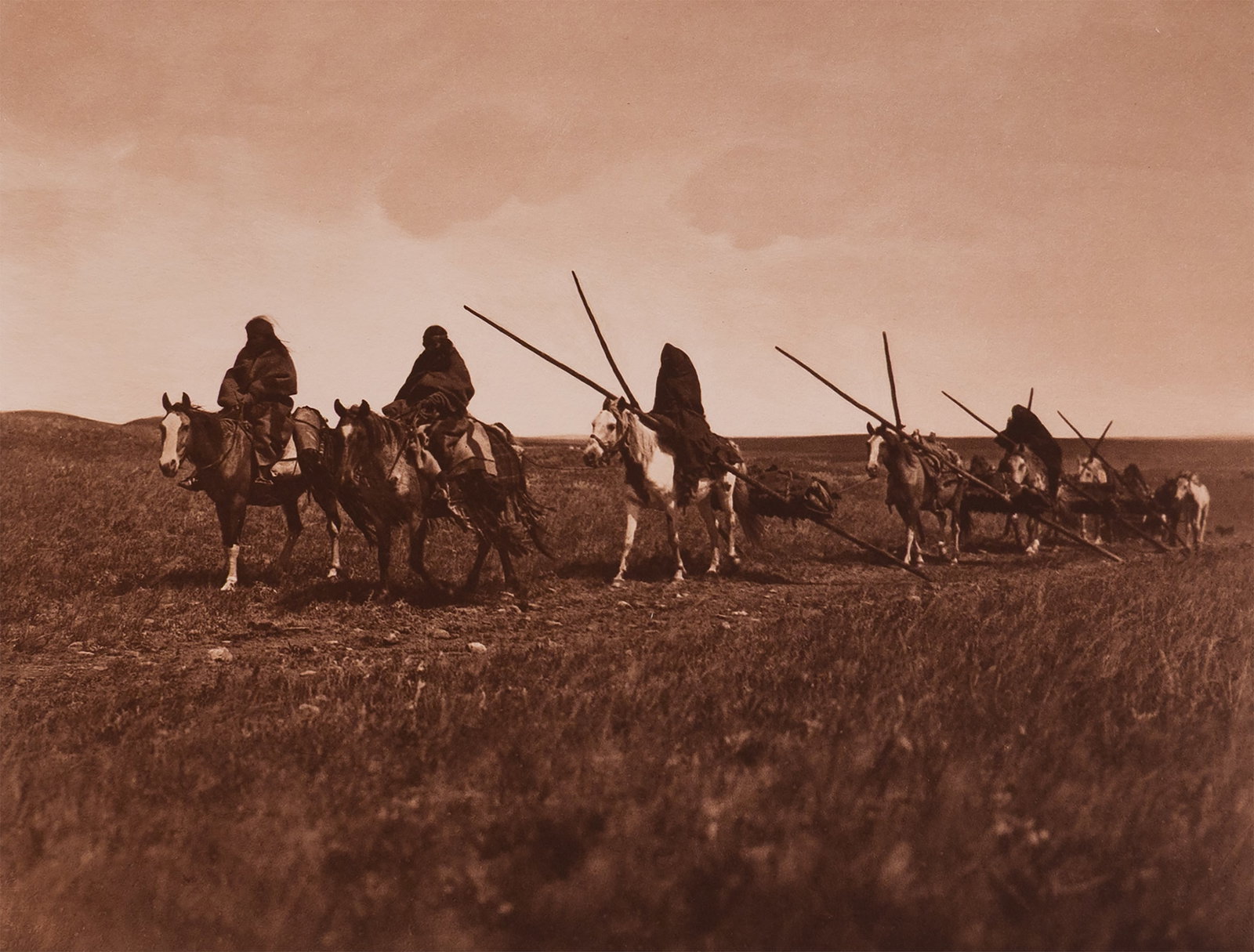 Traveaux - Piegan: This iconic image shows a procession of Piegan women on horseback, each guiding a travois across the prairie. The travois—constructed of two long poles crossed over the horse’s back and dragging b