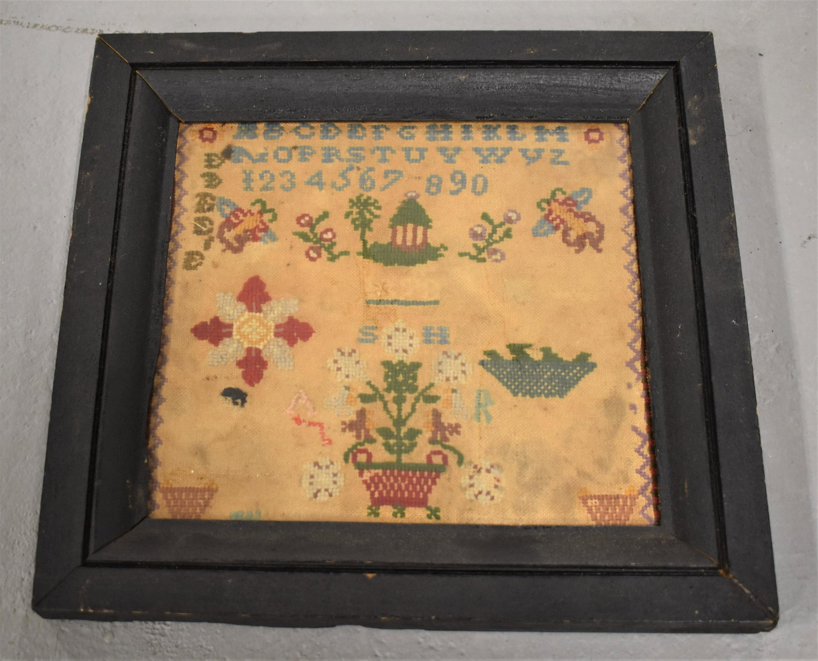 19th Century Needlework Sampler: 19TH CENTURY NEEDLEWORK SAMPLER ON LINEN- ROWS OF LETTERS OVER NUMERALS WITH BASKETS AND FLOWERS IN A BLACK MOLDED WOODEN FRAME- 8 3/4" X 9 3/4" SIGHT- SOILING, STAINS AND IMPERFECTIONS