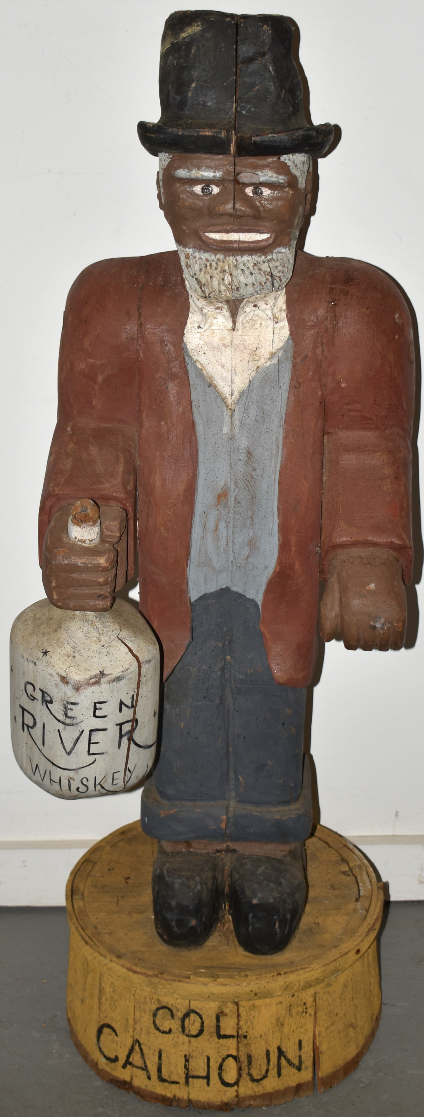 FOLKY CARVED MAN HOLDING GREEN RIVER WHISKEY JUG: 20TH CENT CARVED & PAINTED WOODEN 47" FIGURE OF A BLACK MAN W/ BLACK TALL HAT IN RED JACKET & BLUE VEST HOLDING A GREEN RIVER WHISKEY JUG STANDING ON A BARREL MARKED COL. CALHOUN - SPLITS, WEAR, BOTTO