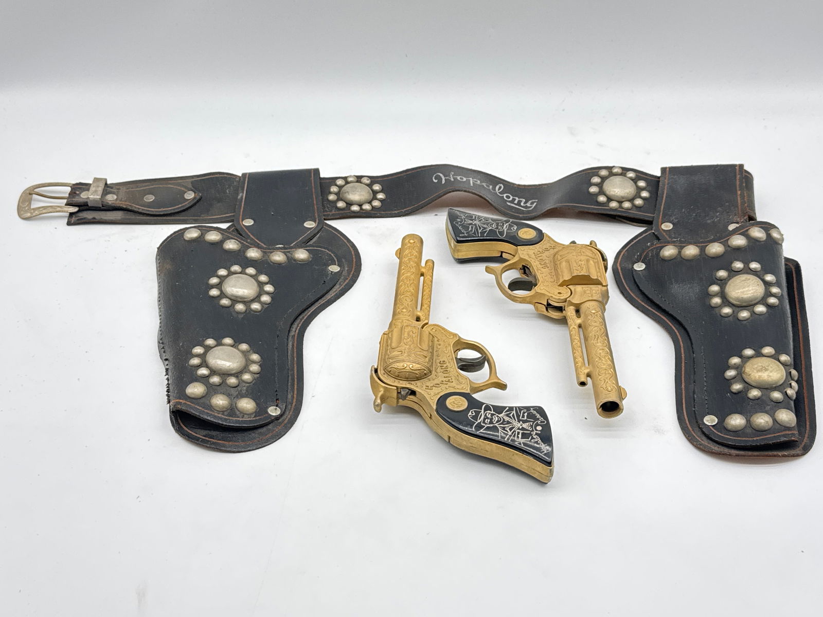 1950s Hopalong Cassidy Double Holster Cap Gun Set by Hubley (147 white): Saddle up with this vintage 1950s Hopalong Cassidy cap gun set! This double holster belt features two matching die-cast metal cap pistols with engraved detailing and black grips bearing Hopalong’s l