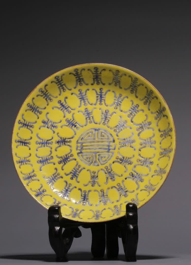 A Qing Dynasty yellow blue color longevity pattern plate: Qing Dynasty yellow ground blue color Wanshou pattern plate open mouth, shallow arc belly, and circle foot below. The outer wall is glazed with white glaze and painted with pastel flower patterns. The