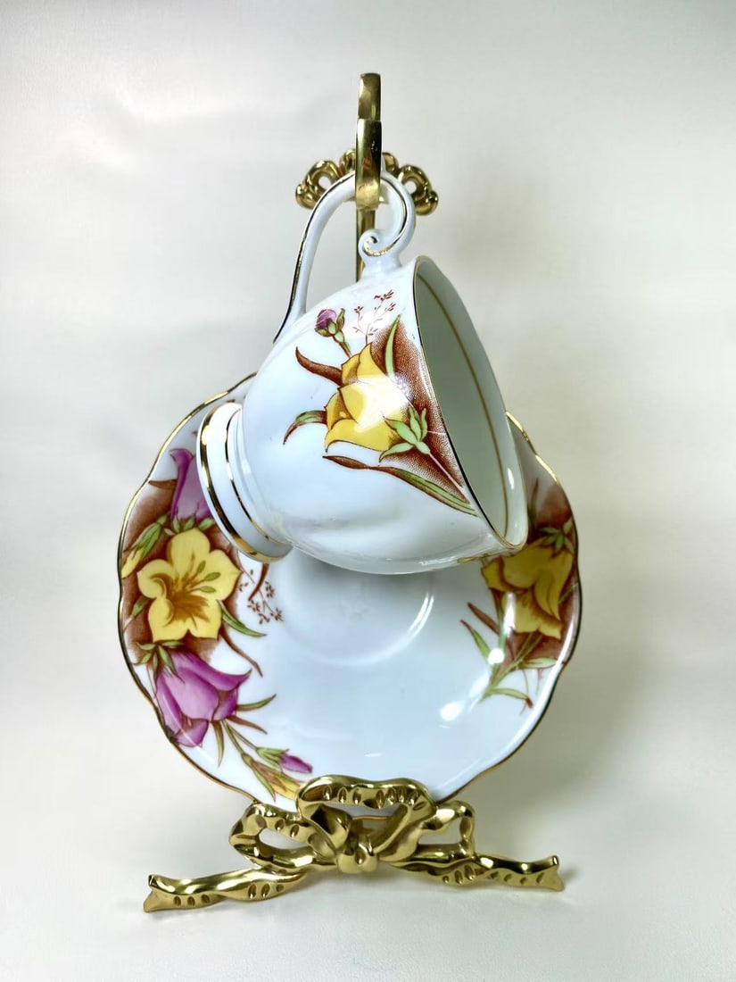 Bell Fine Bone China Floral & Gold Trim Tea Cup and Saucer Set - England: This exquisite tea cup and saucer set by Bell Fine Bone China features a vibrant floral motif in warm yellow and purple hues, accentuated with delicate gold trim. The cup and saucer are adorned with b