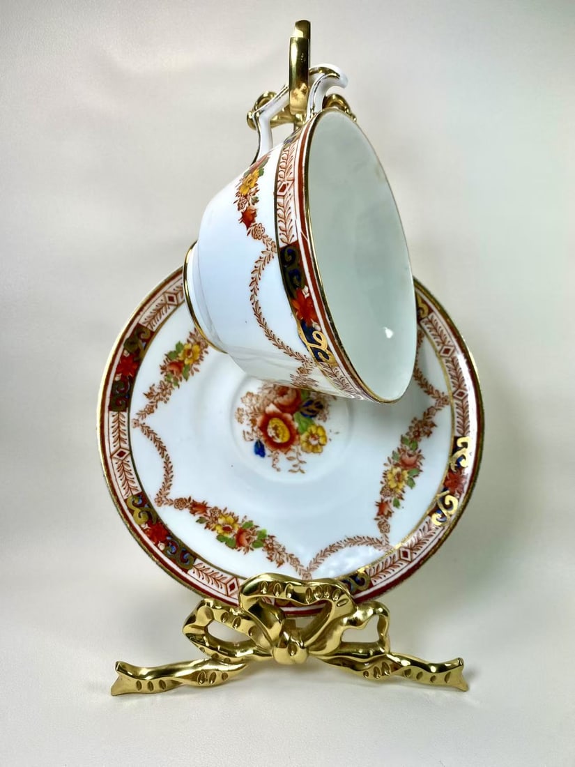 Hanley Hancock's Fine Bone China Floral & Gold Trim Tea Cup and Saucer Set - England: This exquisite fine bone china tea cup and saucer set by Hanley Hancock's features a beautifully detailed floral motif with warm, rich tones, accented by a luxurious gold trim. The ornate handle desig