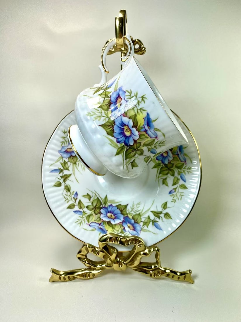 Queens by Rosina China Ltd. Wild Flowers Blue Morning Glory Tea Cup and Saucer Set: A charming tea cup and saucer set by Foley England, adorned with a delicate pastel blue background, intricate gold floral patterns, and a hand-painted flower bouquet at the center. A beautifully craft