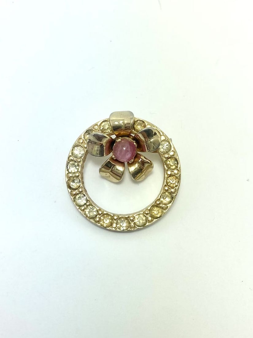 Vintage Coro Gold Tone Rhinestone Circle & Flower Brooch: A vintage brooch by Coro, featuring a gold-tone circular frame set with sparkling rhinestones and a raised floral centerpiece accented with a pink cabochon-style stone. A beautiful piece with timeless