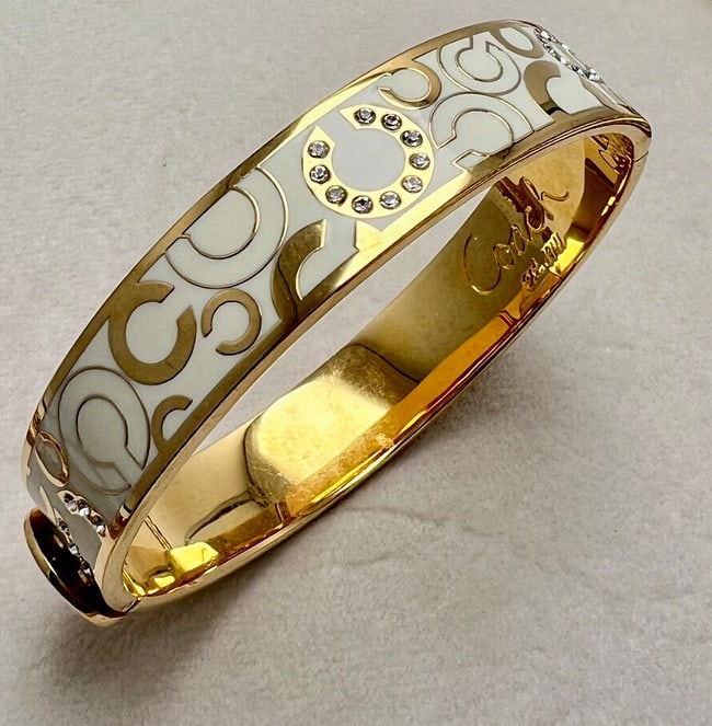 Coach White Enamel Hinged Bangle with Crystal Accents: A contemporary Coach hinged bangle bracelet featuring glossy white enamel with the iconic “C” motif in gold-tone metal. The design is accented with clear round crystals forming stylized “C&r