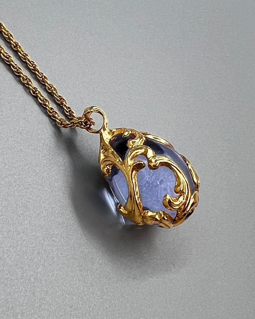 Joan River Gold-Tone Scroll Cage Pendant Necklace with Purple Glass Drop: A vintage necklace by Joan River, featuring a dramatic drop pendant with a faceted purple glass cabochon encased in a gold-tone openwork scroll cage. The elegant design combines a rich amethyst-like h