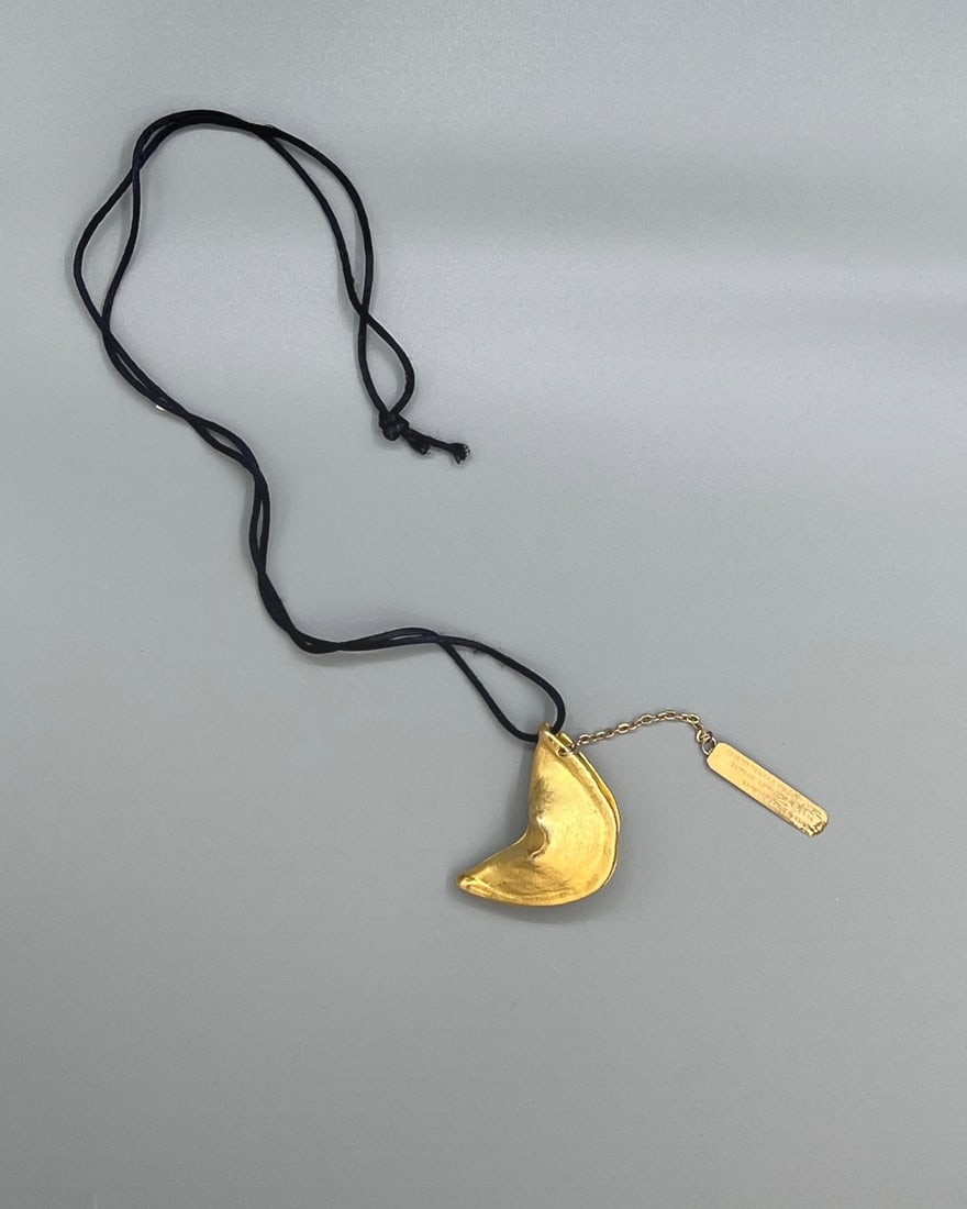 Miriam Haskell Vintage Fortune Cookie Pendant Necklace with Engraved Tag: A whimsical vintage pendant necklace by Miriam Haskell, featuring a sculptural fortune cookie charm in antique gold-tone finish. The textured pendant hangs from a black cord and is accompanied by a da