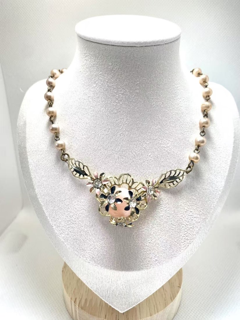 Vintage Pink Moonglow Lucite Floral Necklace with Faux Pearls and Enamel Accents: Charming mid-century floral necklace featuring a central pink moonglow lucite cabochon with a soft, luminous glow, framed by openwork gold-tone petals. The design is accented with small pink and black