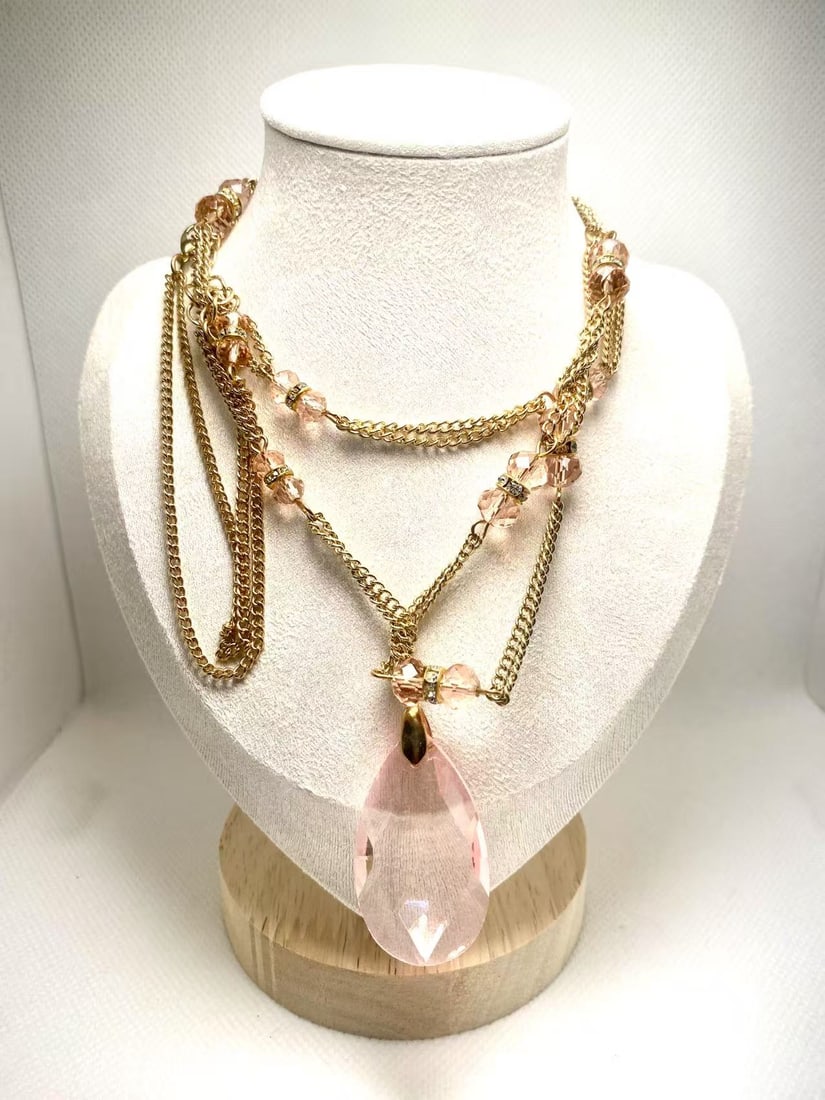 Gold-Tone Chain Long Necklace with Pink Crystal: and small round accents, creating a rich and vintage-inspired look. The focal pendant is a large teardrop-shaped pink crystal with sharp faceting that reflects light beautifully. The chain is embellis