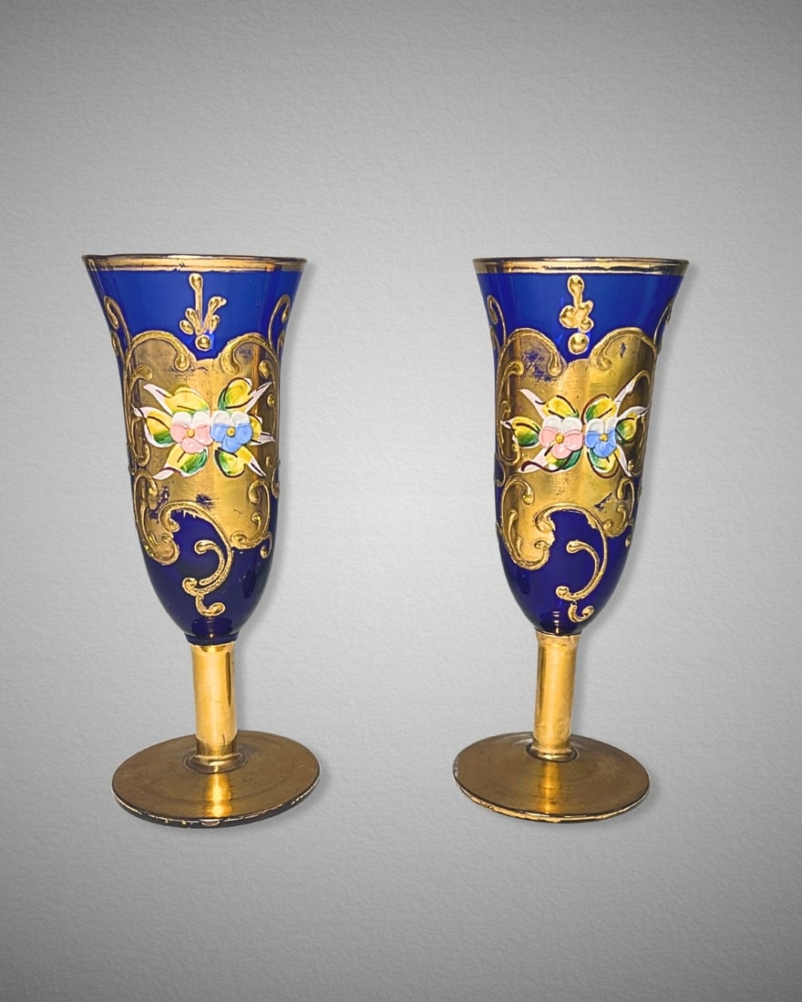 Pair of Mid-Century Bohemian Cobalt Blue Enamelled Glass Goblets: This elegant pair of mid-century Bohemian goblets features deep cobalt blue blown glass with ornate 24K gold enameled scrollwork and hand-applied raised floral accents in pink, yellow, and blue. The t