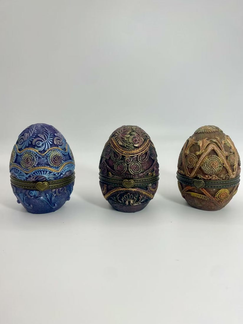 Set of 3 Decorative Egg Trinket Boxes _ Faberg?? Style: A set of three decorative egg-shaped trinket boxes with ornate embossed designs and antique-style heart clasps. Featuring rich colors of blue, purple, and bronze tones, these Faberg??-inspired eggs op