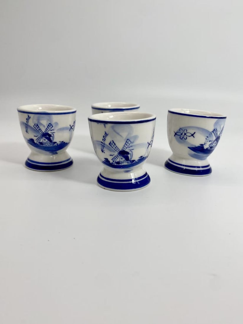 Lot of 4 Hand-Painted Delft Style Blue & White Egg Cups _ Holland: This lot offers a set of four hand-painted Delft style egg cups, decorated in blue-and-white with traditional windmill and floral motifs. Each piece is marked ??Handpainted Blue Holland,?? highlightin