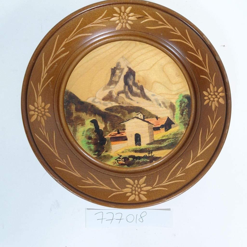 Vintage Painted Wood Plate - Alpine Mountain: Vintage decorative wooden plate featuring a hand-painted alpine mountain landscape with village scene, framed by carved floral motifs. Turned wood construction with raised footed base. Pre-owned condi