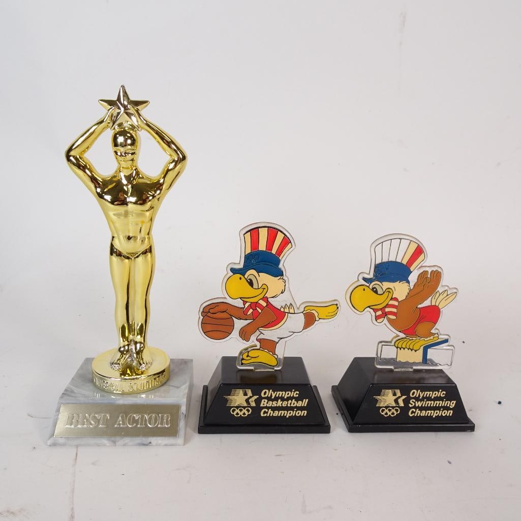 Vintage Trophy Lot (3): Vintage lot of three collectible trophies, featuring one gold-tone Universal Studios "Best Actor" statue and two 1984 Olympic-themed eagle character trophies for Basketball and Swimming. All pieces sh