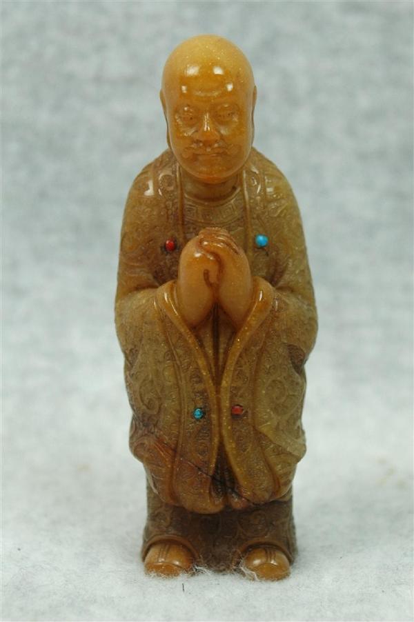 Chinese Tian-Huang Stone Figurine: Chinese tianhuang stone figurine, well carved and incised as a louhan (Buddhist sage) standing in elaborate brocade monastic robes with shou medallions amid scrolling leafy tendrils, his hands held to