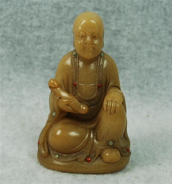 Chinese Tian-Huang Stone Figurine: Chinese tianhuang stone figurine of a Buddhist sage, the smiling louhan seated in monastic robes and holding a wish-granting scepter in his right hand, the rich yellow matrix with inlaid green and red