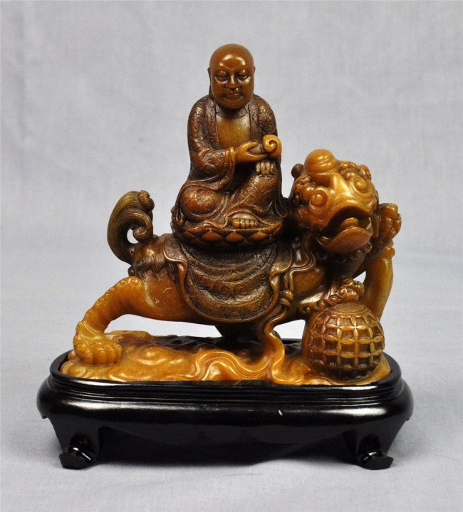 Chinese shoushan stone figural carving: Chinese shoushan stone figural carving, featuring a Buddhist sage seated in brocade patterned robes and holding a scepter to the front while riding on the back of a fu-lion playing with a large ball, 
