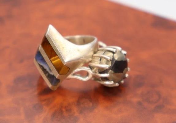 Two sterling silver rings: One featuring a geometric inlay of various stones including tiger's eye, and the other a round, possibly moonstone or similar pale gemstone, set in a claw-style setting. Boxes not included. 