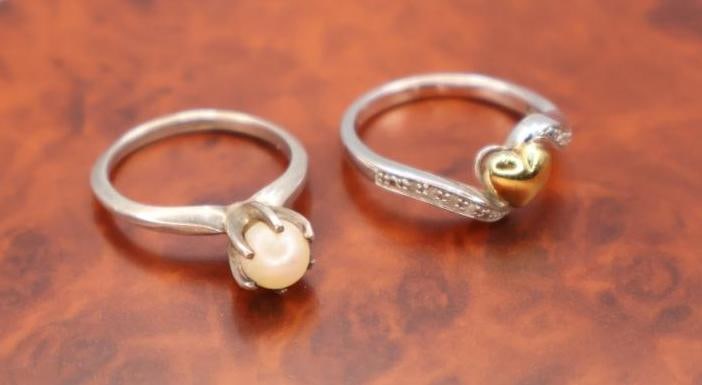 A pair of sterling silver rings: One featuring a white round pearl in a prong setting, and the other a wave design with a gold-toned heart accent and small paved stones. 