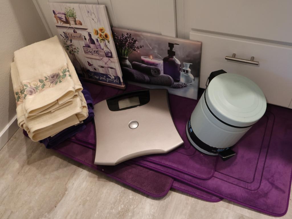 Bathroom Essentials Including Mats: Purple bathrooms mats, small bathroom trash, decorative bathroom towels, two pieces of art, and a bathroom scale. 