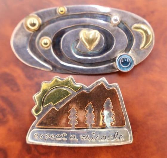 Two Silver Pins with Metal Accents: Orbiting heart pin with blue glass accent, and 'Expect and Miracle' mountain pin