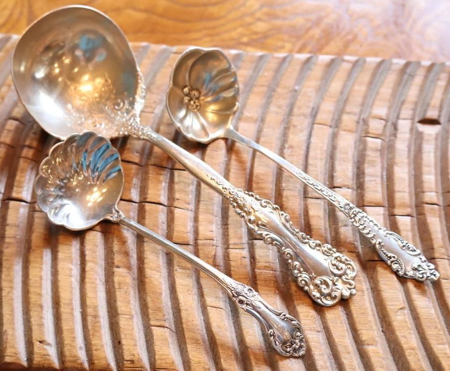 Three Sterling Silver Ornate Serving Spoons, 125 Grams: Three ornate silver serving spoons in assorted patterns, including shell-form and floral-decorated bowls. Sterling marks visible on multiple pieces. Combined weight: 125 grams. Vintage condition with
