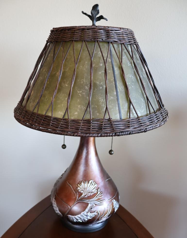 Quoizel Highland 2 Light Table Lamp: Features an organic, soft green oyster mica shade wrapped in a woven wicker overlay. Typically stands around 25 inches tall with a 16.5-inch shade diameter. Topped with a sculpted pine leaf/twig finia