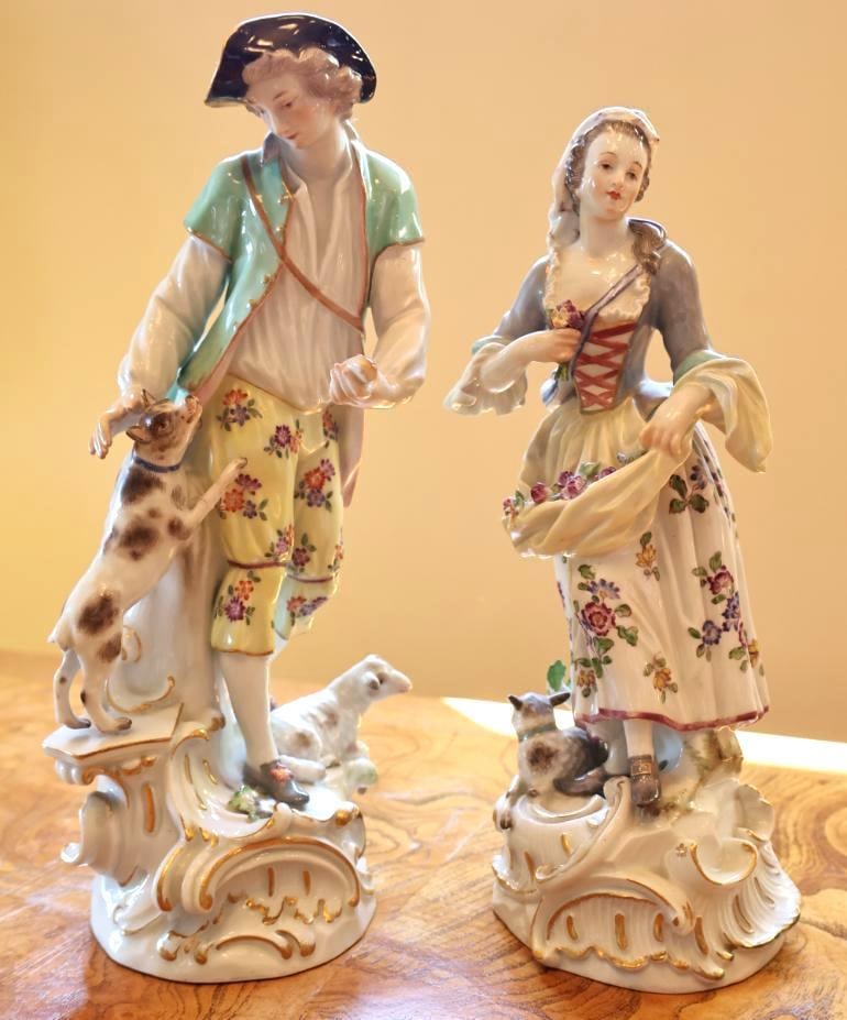 Pair of Meissen Porcelain Pastoral Figurines: Pair of decorative porcelain pastoral figurines depicting an elegantly dressed woman with flowers and a gentleman accompanied by a dog and lamb. Damage is present, including a broken/missing applied