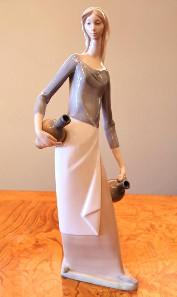 NAO by Lladro Woman Carrying Water Jugs Porcelain Figurine: Finished in soft gray, cream and brown tones with the elongated styling characteristic of NAO figures. Made in Spain and marked on underside. Measures approximately 13" H.