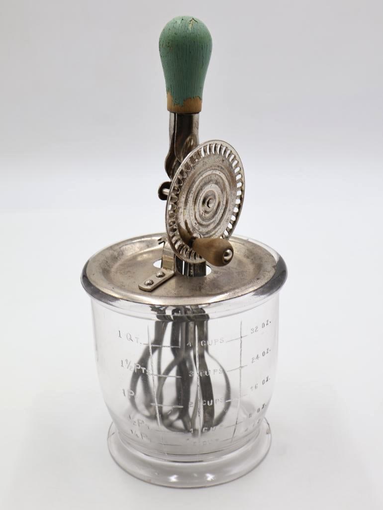 Vintage Hand-Crank Egg Beater with Glass Measuring Jar: Vintage hand-crank rotary egg beater and kitchen mixer featuring a clear glass measuring jar with graduated cup and ounce markings, metal lid and gear mechanism, and a painted turquoise wooden