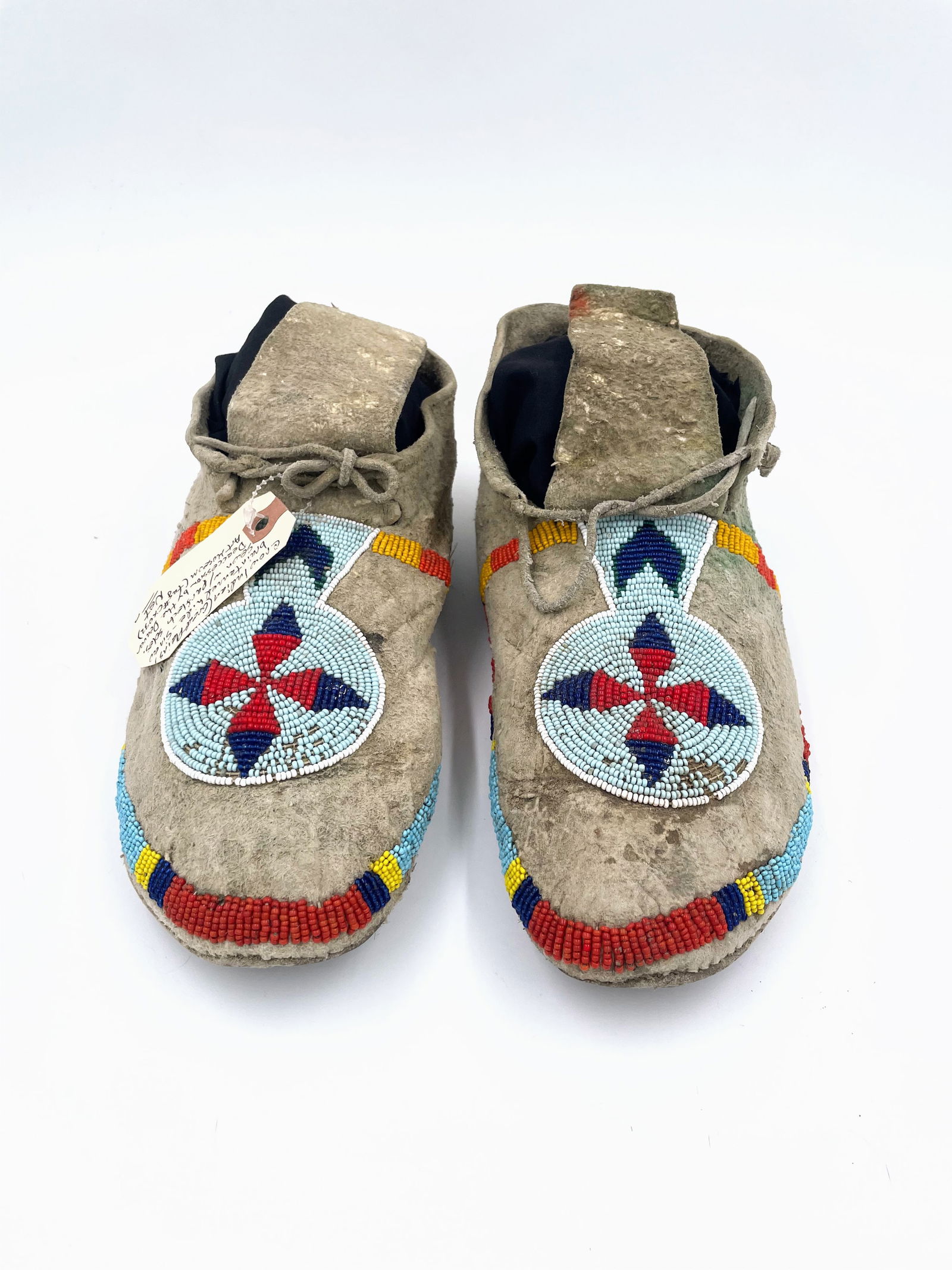 Crow Beaded Moccasins (Turn of the Century): Finely crafted hide moccasins from the Crow Nation, featuring exquisite turn-of-the-century beadwork in classic concentric cross and chevron patterns. Executed in red, sky blue, navy, yellow, and whit