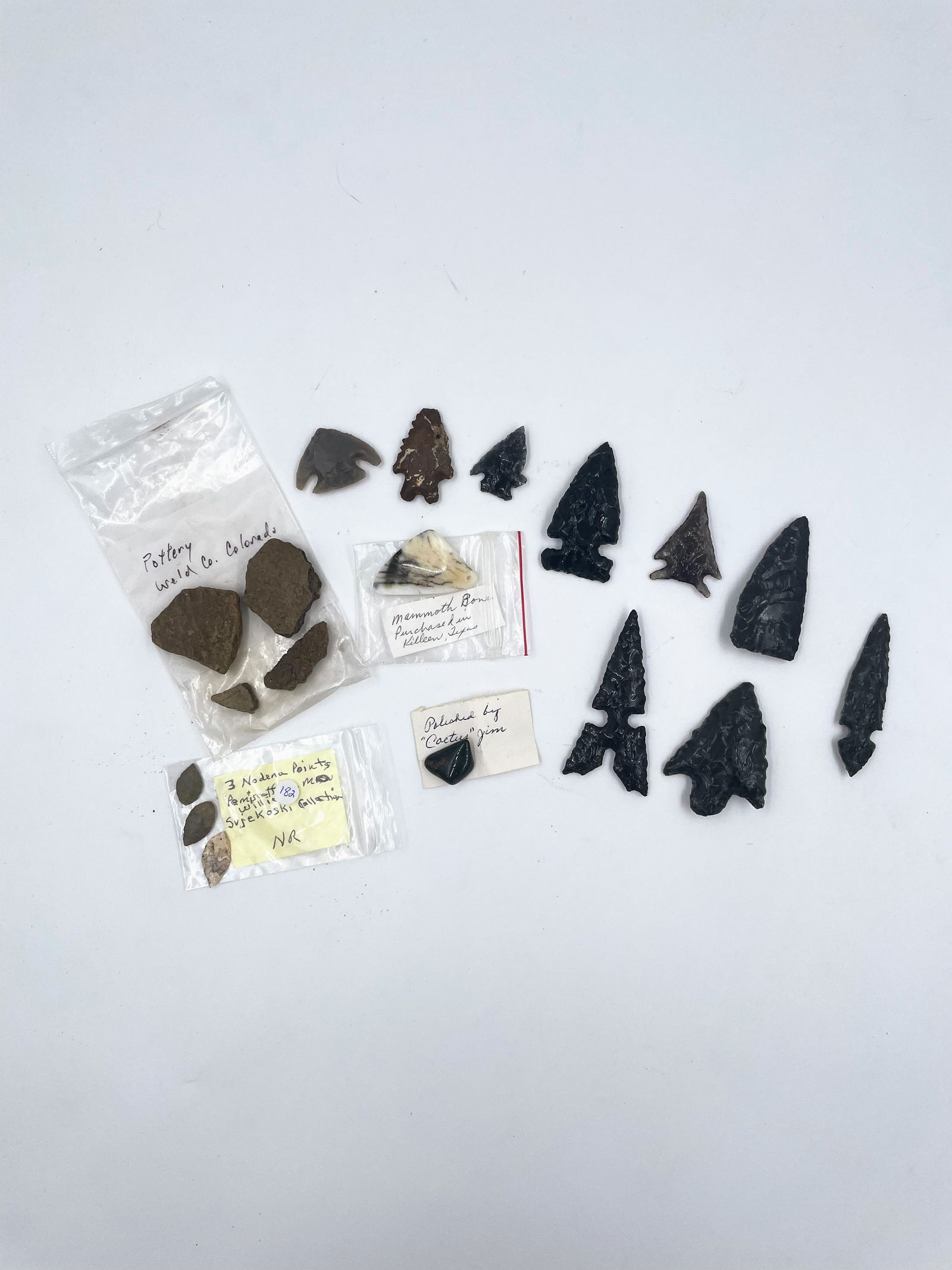 Group of Arrowheads: A varied assortment of stone artifacts, including several finely knapped obsidian points, along with labeled fragments of pottery, bone, and additional lithics. This grouping spans a mix of materials