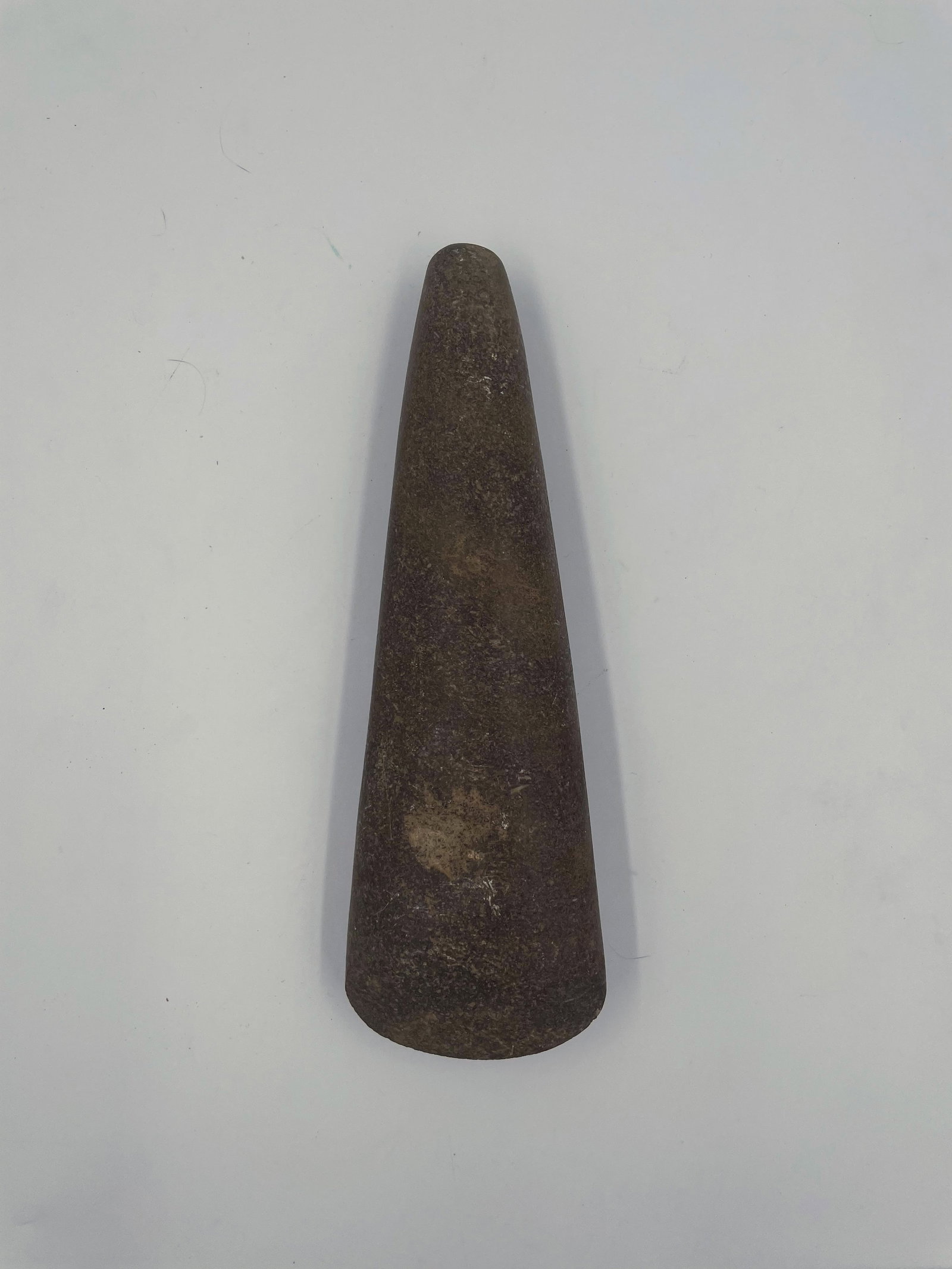 Celt: Offered here is a Native American celt, carefully shaped and ground from dense stone. The piece features a tapered, symmetrical form with a rounded cutting edge and smoothed surface, measuring just ov