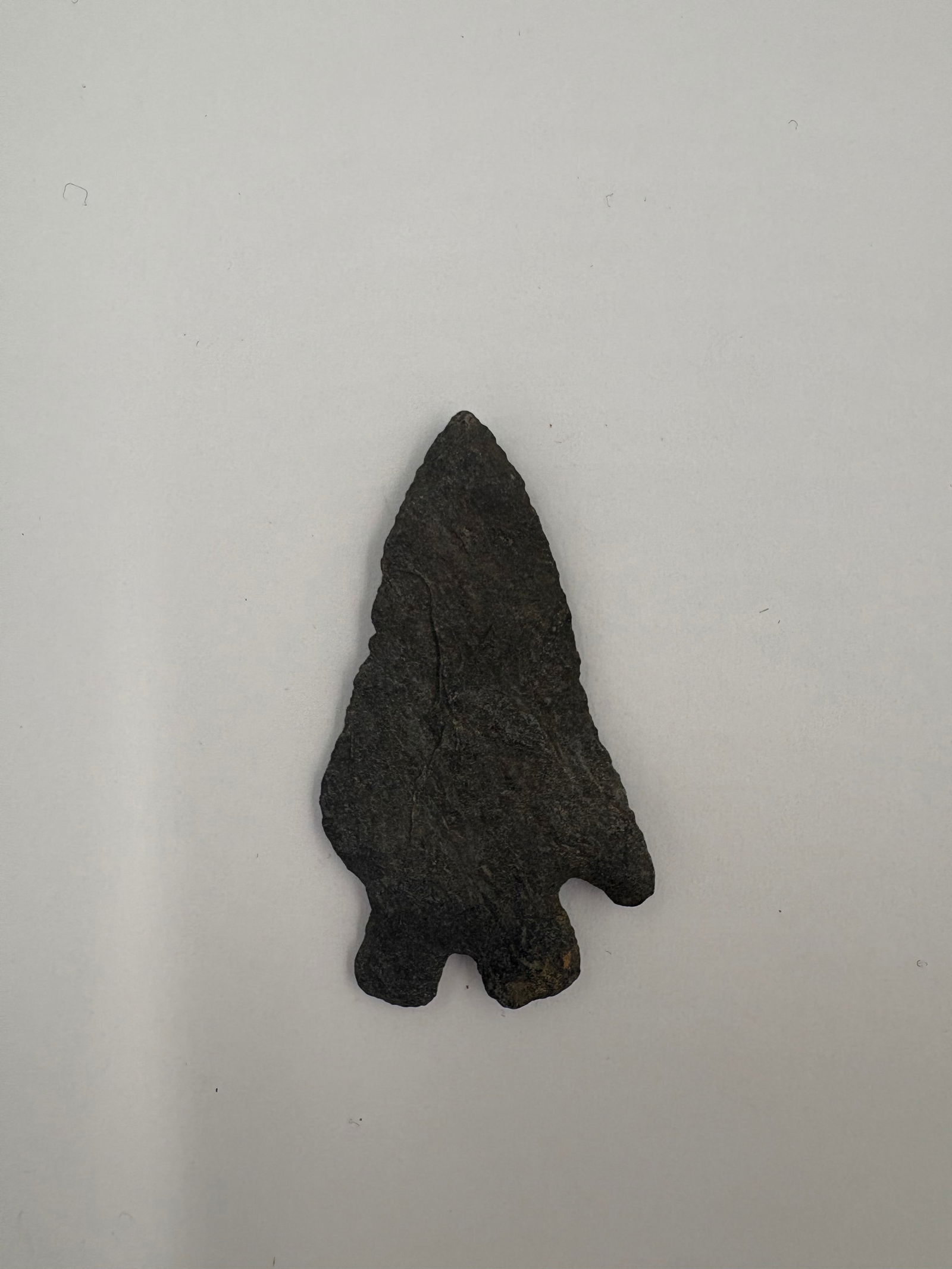 Kirk - 2in: Dark gray to black stone projectile point with a narrow triangular blade and bifacial flaking along the edges. Pronounced basal notches form a three-lobed base and a darker surface patina is present a