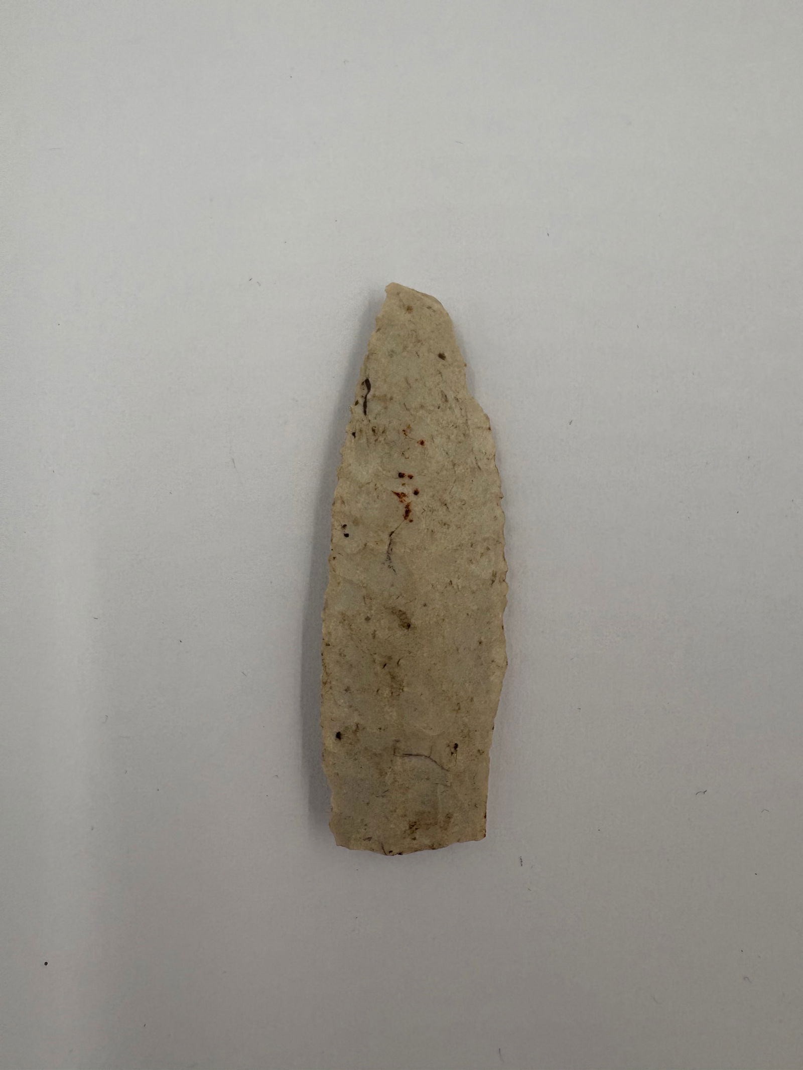 First View - 2 1/4in: Elongated flaked stone blade measuring 2 1/4 in with a well-defined median ridge running along its length. Edges display oblique flaking that produces a narrow, tapered profile and fine micro-flake