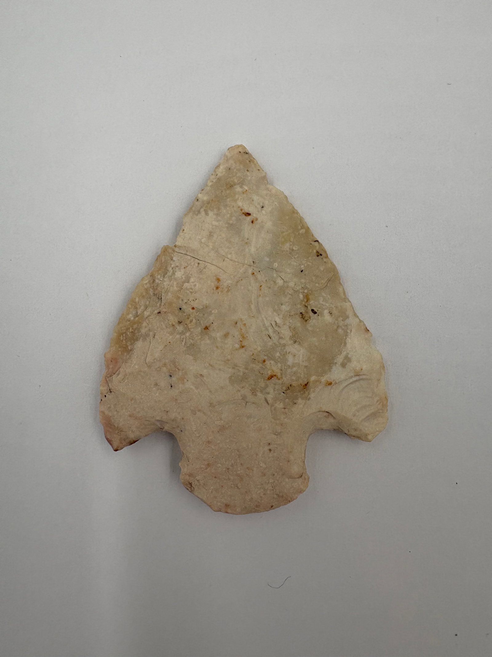 Snyder: Beige to light-tan stone projectile point with a broad triangular blade and pronounced notched basal ears. Flake scars and a subtle patina cover both faces, with small mineral spots and slight edge ir