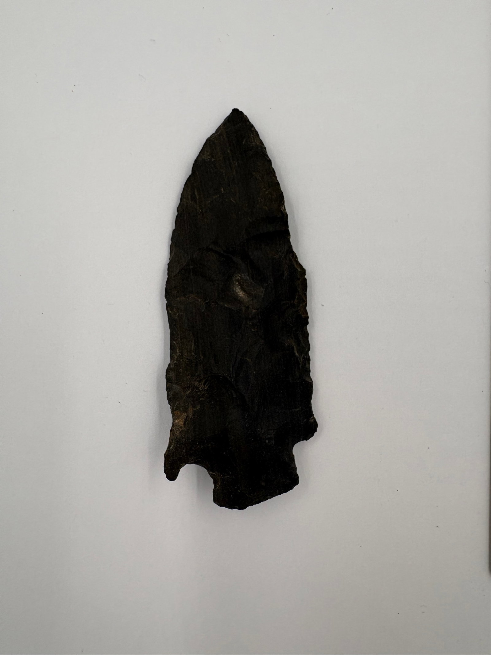 Etley: Dark stone projectile point featuring a slender, tapered blade and distinct notched basal ears with bifacial flaking evident on both faces. Matte black coloration with prominent flake scars and a slig