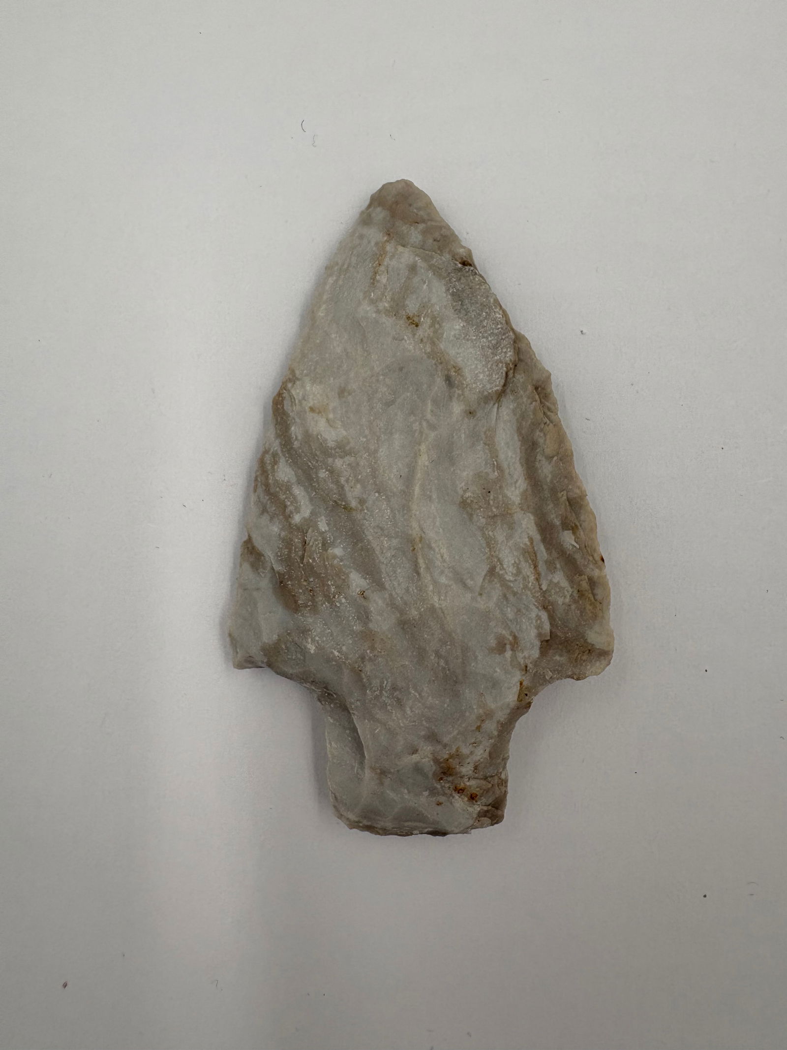 Smith: Light gray hand-flaked stone projectile point with a triangular, bifacially worked blade and subtle side notches leading to a narrow stemmed base. Fine flake scars cover the surface and faint brown mi